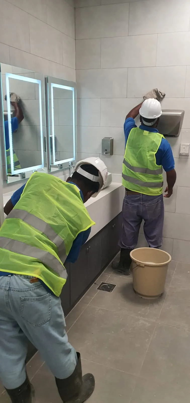 Two workers cleaning the mirrors, tiled wall and vanity in a washroom
