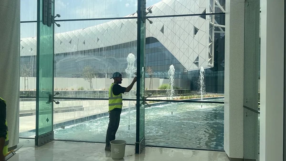 عامل يمسح جدارًا زجاجيًا تظهر عليه آثار الماء أمام نافورة