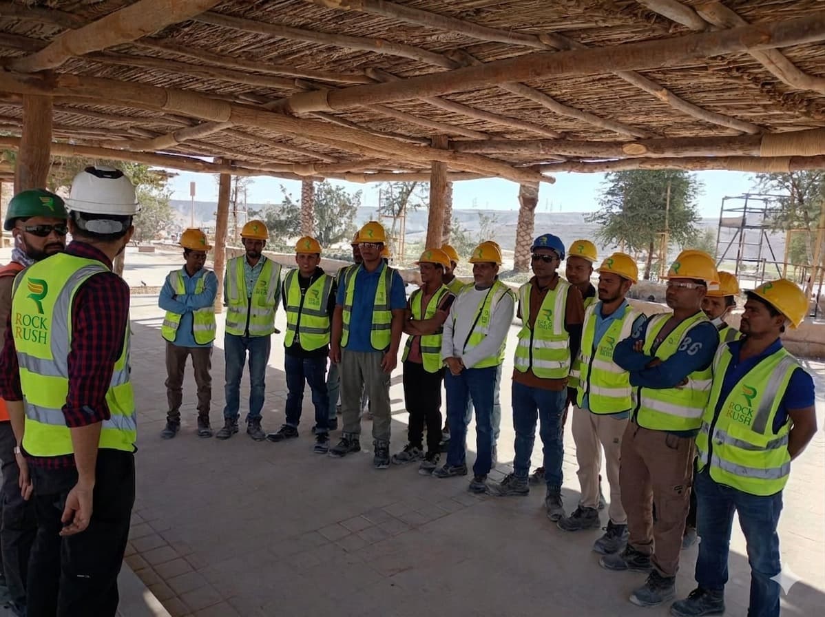 طاقم روك راش من سبعة فنيين بقمصان العلامة الخضراء وشعار R الكهرماني، يحمل كلٌّ منهم أداة احترافية — مثقاب لاسلكي، ومَمْسحة زجاج، ورذاذ كهروستاتيكي، ومنظف بخاري، ومكنسة تجارية، وعدّادات تكييف، وشفاط ورش — يقفون أمام برج مكاتب حديث في الرياض