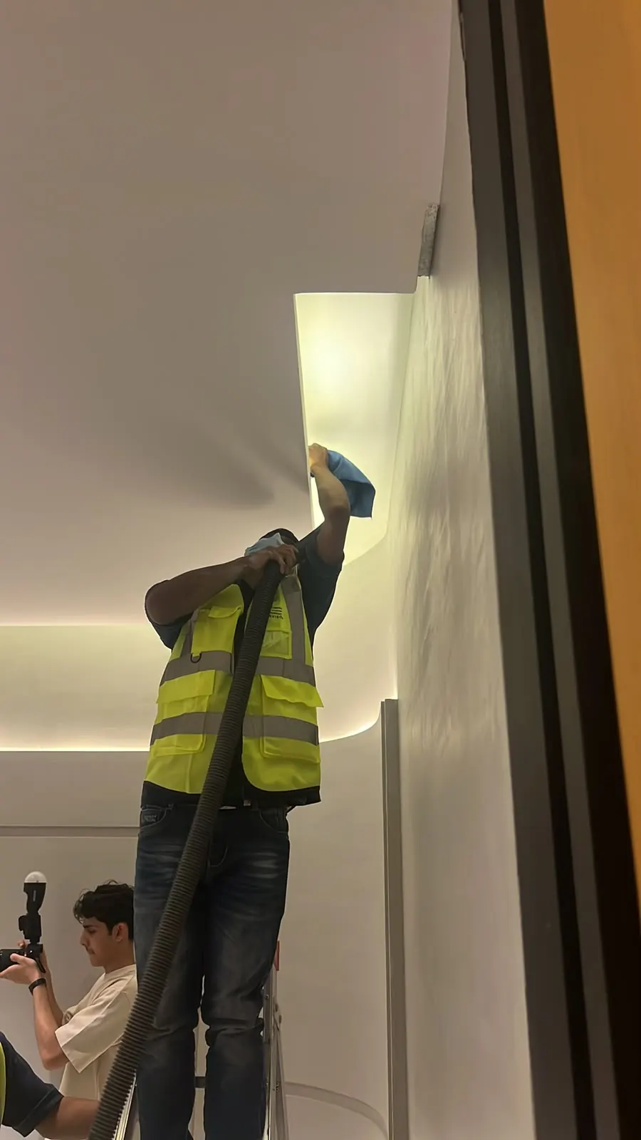 A worker on a ladder vacuuming and wiping a ceiling cove ledge